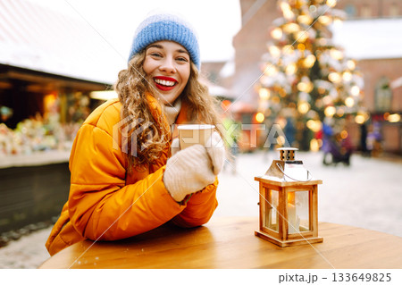 A woman in a bright jacket and red lips walks through a Christmas market and drinks a hot drink. 133649825