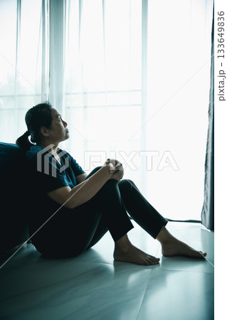 A lonely woman sits on the floor by window looking outside in dark room. Concept of depression, anxiety, sadness, loneliness, mental health problem, emotional struggle and hope for better life. 133649836