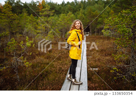 Hiker woman with a backpack hiking on a wooden path in a swamp forest. Active lifestyle Hiker woman with a backpack hiking on a wooden path in a swamp forest. Active lifestyle 133650354