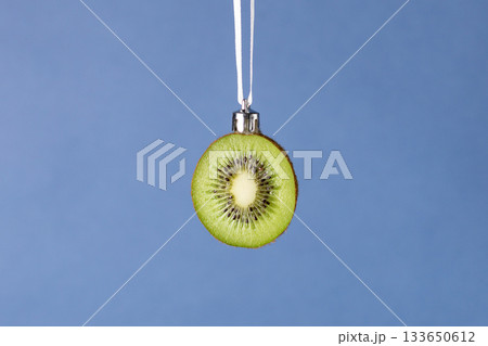 Creative christmas ornament: fresh kiwi slice hanging on white ribbon on blue background Creative christmas ornament: fresh kiwi slice hanging on white ribbon on blue background 133650612