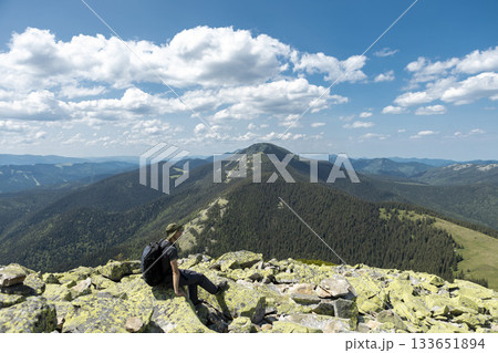 A tourist with a backpack and hat in the mountains sits on a rock and looks into the distance, a summer cloudy day A tourist with a backpack and hat in the mountains sits on a rock and looks into the distance, a summer cloudy day 133651894