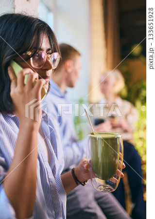 Young woman holding matcha and talking on phone in relaxed office environment 133651928