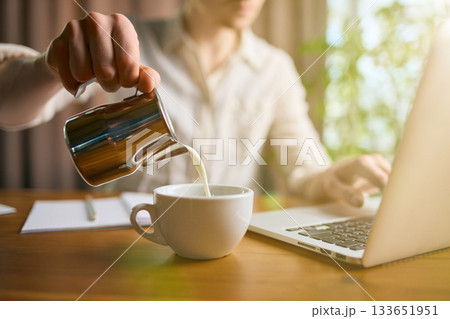 Man pouring milk into coffee cup while working on laptop Man pouring milk into coffee cup while working on laptop 133651951