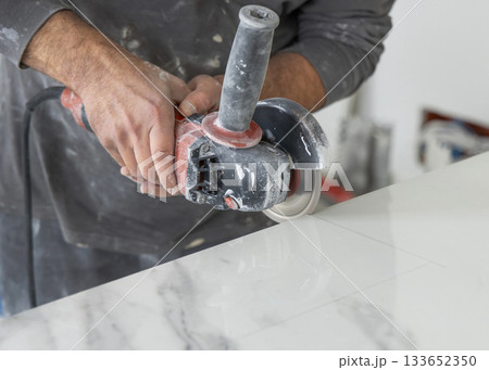 Craftman in grey shirt cuts a gray marble tile using an angle grinder. Handy men at work, hands closeup Craftman in grey shirt cuts a gray marble tile using an angle grinder. Handy men at work, hands closeup 133652350