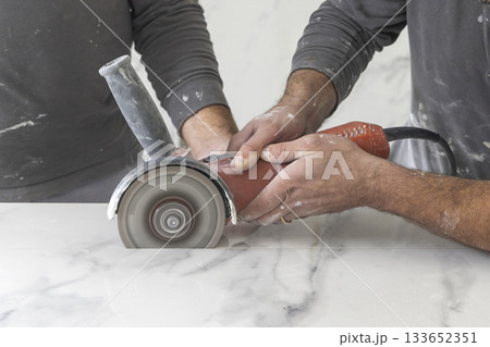 Craftman in grey shirt cuts a gray marble tile using an angle grinder. Handy men at work, hands closeup Craftman in grey shirt cuts a gray marble tile using an angle grinder. Handy men at work, hands closeup 133652351