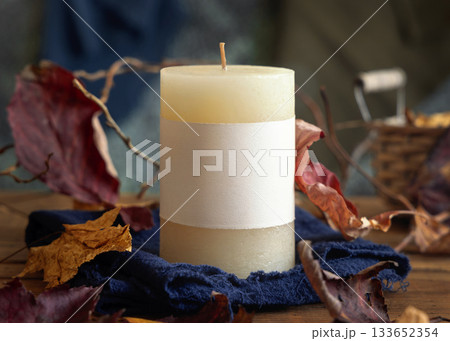 Candle with a label near autumn dried leaves closeup,  fall mockup in blue, red and orange 133652354