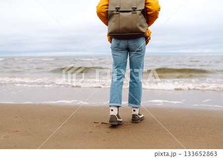 Woman traveler walks in boots along the seashore. Happy traveler with a backpack enjoys the sea air. Woman traveler walks in boots along the seashore. Happy traveler with a backpack enjoys the sea air. 133652863