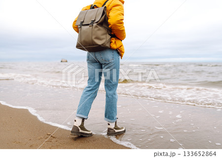 Woman traveler walks in boots along the seashore. Happy traveler with a backpack enjoys the sea air. 133652864