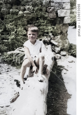 A vintage portrait of a boy sitting on a log near an ancient stone wall. Retro photo from 1965. 133655098