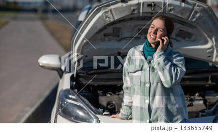 Caucasian woman laughing and calling on phone asking for help standing near car with open hood.  133655749