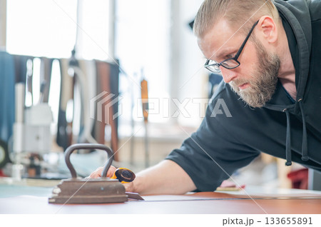 Leatherworker measuring and cutting leather in workshop. Leatherworker measuring and cutting leather in workshop. 133655891