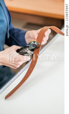 A woman makes holes in a leather belt. Leatherworker's workshop. A woman makes holes in a leather belt. Leatherworker's workshop. 133655947