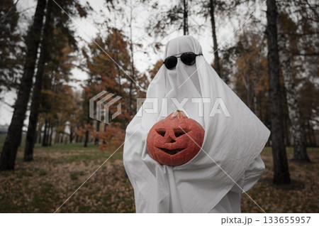 Woman in white sheet holding jack o'lantern against autumn forest background. Halloween costume. Woman in white sheet holding jack o'lantern against autumn forest background. Halloween costume. 133655957
