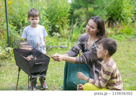 Caucasian woman grilling sausages on a barbecue with her two sons. Caucasian woman grilling sausages on a barbecue with her two sons. 133656302