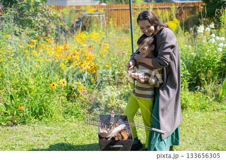 Caucasian woman grilling sausages on a barbecue with her son. Caucasian woman grilling sausages on a barbecue with her son. 133656305