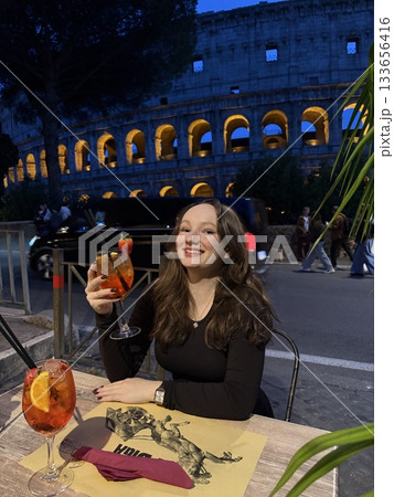 Rome, Italy 11.29.2025 Aperol in front of the Colosseum girl sitting in a restaurant near the Colosseum, eating pizza in evening, people walking in background, talking, blogging, and seeing sights. 133656416