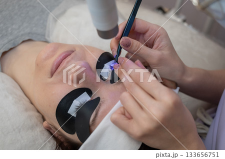 A woman undergoing eyelash extension procedure using an ultraviolet lamp. A woman undergoing eyelash extension procedure using an ultraviolet lamp. 133656751