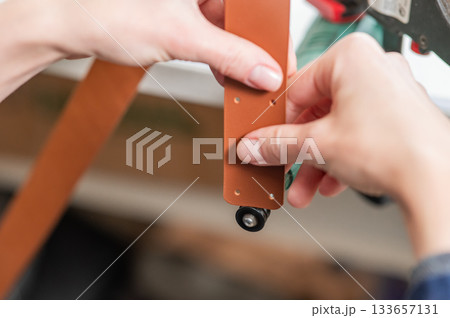 Leatherworker grinding the edge of a strip of leather for a belt in the workshop.  133657131