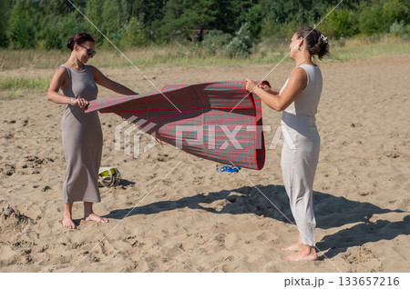 Two Caucasian women spread a blanket on the beach for a picnic. Two Caucasian women spread a blanket on the beach for a picnic. 133657216