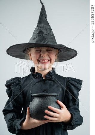 Portrait of a little Caucasian girl in a witch costume holding a cauldron on a white background. Vertical photo. Portrait of a little Caucasian girl in a witch costume holding a cauldron on a white background. Vertical photo. 133657287