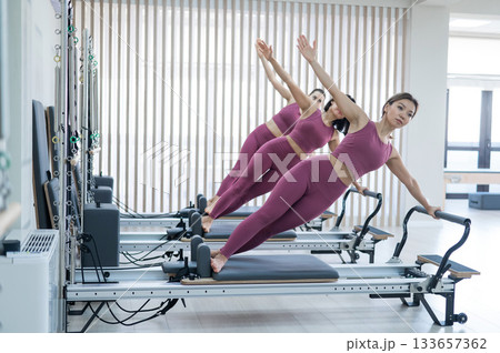 Three Asian women in pink sportswear doing side planks on a reformer. Pilates classes. Three Asian women in pink sportswear doing side planks on a reformer. Pilates classes. 133657362