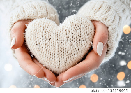 Soft white knitted heart held in hands against a snowy background with softly glowing lights during winter season Soft white knitted heart held in hands against a snowy background with softly glowing lights during winter season 133659965