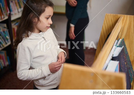 Young Girl Exploring Books in School Library While a Teacher Oversees Learning Young Girl Exploring Books in School Library While a Teacher Oversees Learning 133660583