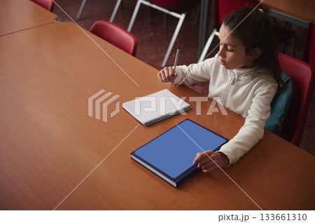 Young Girl Studying At A Table With Notebook And Blue Book In Classroom Setting Young Girl Studying At A Table With Notebook And Blue Book In Classroom Setting 133661310