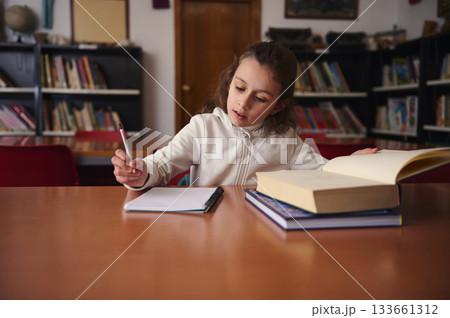 Young Girl Studying In Library With Open Book, Notebook, And Large Textbooks Nearby Young Girl Studying In Library With Open Book, Notebook, And Large Textbooks Nearby 133661312