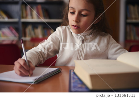 Focused Girl Writing in Notebook at Library Desk with Open Books Nearby Focused Girl Writing in Notebook at Library Desk with Open Books Nearby 133661313