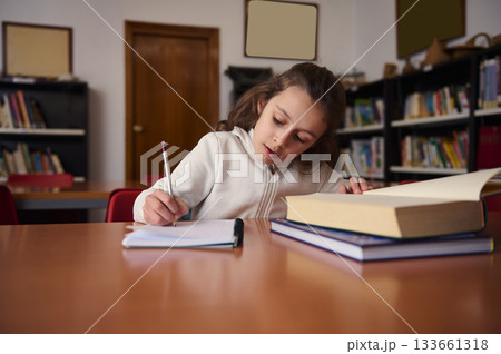 Focused Child Studying in Library, Writing in Notebook Beside Stack of Books Focused Child Studying in Library, Writing in Notebook Beside Stack of Books 133661318