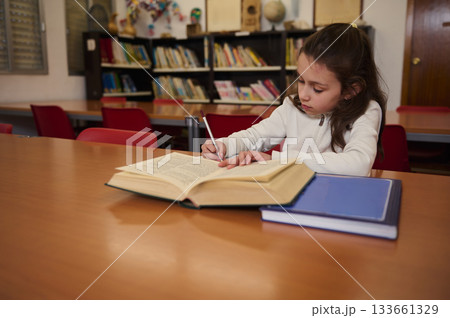 Young Girl Studying at Library Table, Writing in a Large Open Book, Focused and Thoughtful 133661329