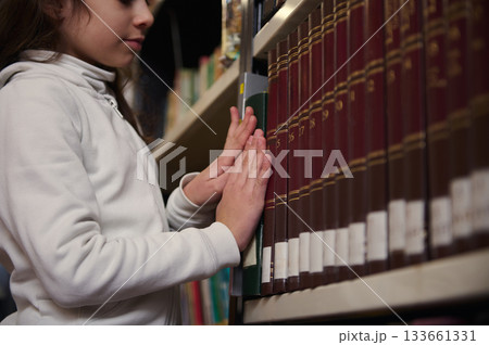 Young Girl In White Hoodie Exploring Books On A Library Shelf 133661331