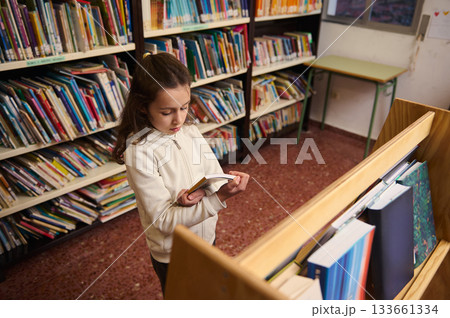 Little Girl Reading a Book in a Public School Library for Book Day Celebration Little Girl Reading a Book in a Public School Library for Book Day Celebration 133661334