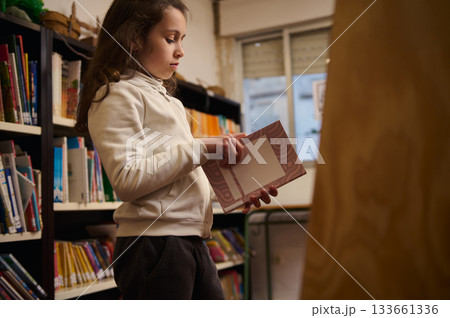 Young Girl Reading a Book in a Public Library, Quiet Focused Moment Among Shelves Young Girl Reading a Book in a Public Library, Quiet Focused Moment Among Shelves 133661336