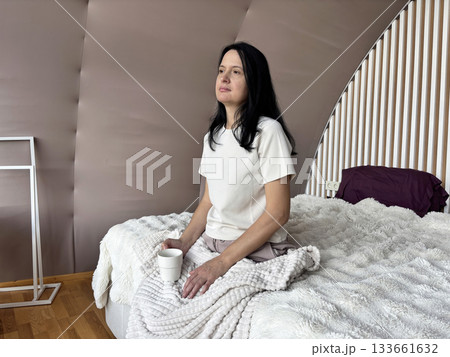 Calm adult woman sitting on bed with warm cup of coffee in relaxed morning atmosphere, peaceful home routine, comfort and wellness lifestyle concept Calm adult woman sitting on bed with warm cup of coffee in relaxed morning atmosphere, peaceful home routine, comfort and wellness lifestyle concept 133661632