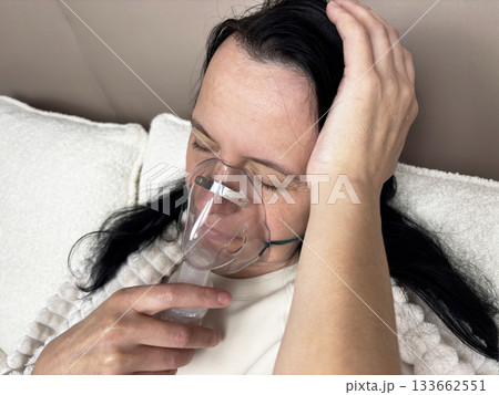 Adult brunette woman inhaling medication through a portable nebulizer device in a bedroom environment, home respiratory care, chronic illness management, realistic medical lifestyle photo 133662551