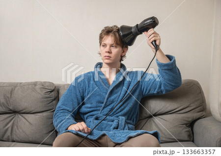 Young caucasian man in blue bathrobe using black hairdryer to dry his hair while sitting on sofa. Mens Home Self-Care Routine Young caucasian man in blue bathrobe using black hairdryer to dry his hair while sitting on sofa. Mens Home Self-Care Routine 133663357