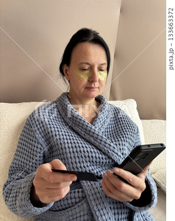 Woman sitting at home holding a bank card and smartphone, making secure online payment, mobile banking, shopping from home, digital finance and modern technology lifestyle concept 133663372