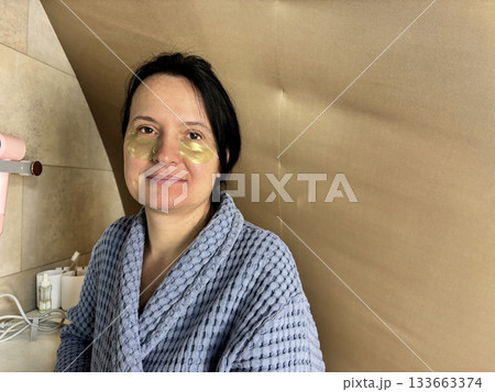 Relaxed woman applying under eye patches during morning skincare routine in a bright home bathroom, self care ritual, facial treatment, natural beauty and wellness lifestyle concept 133663374