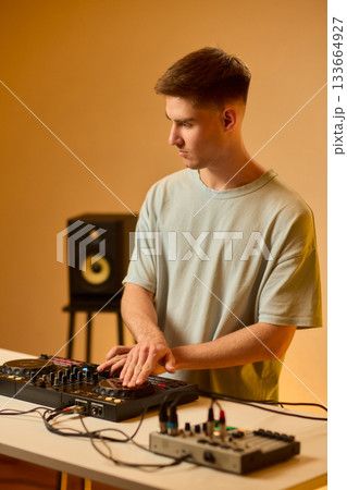 Young DJ with headphones, Skilled DJ engaging with equipment in lively environment, Energetic DJ prepping for live performance amid colorful lighting and fashionable attire 133664927
