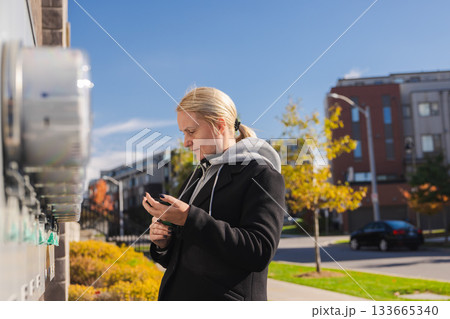 Middle-aged woman reading outdoor electricity meters and entering usage data on smartphone in a residential area on a sunny autumn day Middle-aged woman reading outdoor electricity meters and entering usage data on smartphone in a residential area on a sunny autumn day 133665340