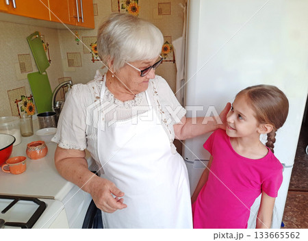 A grandmother with glasses and a white apron in the kitchen with her granddaughter. A grandmother with glasses and a white apron in the kitchen with her granddaughter. 133665562