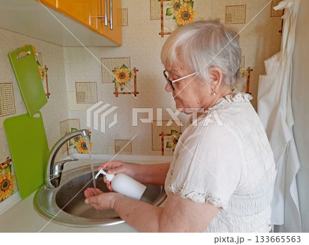 An elderly woman washes her hands with soap under running water from a faucet in a kitchen sink. An elderly woman washes her hands with soap under running water from a faucet in a kitchen sink. 133665563