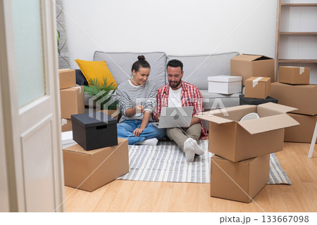 A couple, amidst boxes in their new home, utilizes a laptop for interior design choices, online shopping, or moving arrangements 133667098