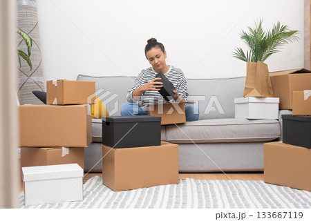 Relocation takes center stage as a young woman sits on the sofa in her new home, unpacking boxes filled with belongings on moving day, emphasizing the real estate experience. 133667119