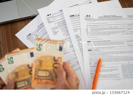 Close-up of hands holding euro bills over various tax forms including W-4, 1040, W-9, and 1120. Tax season, financial accounting, and tax refund concept Close-up of hands holding euro bills over various tax forms including W-4, 1040, W-9, and 1120. Tax season, financial accounting, and tax refund concept 133667124