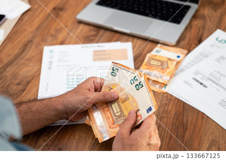 Close-up of hands holding euro banknotes over utility bills and laptop on wooden desk. Concept of personal finance, managing household expenses, and budgeting  133667125