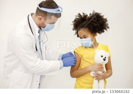 Male doctor in white medical gown stick a band-aid after vaccination. Man wearing white medical gown and face mask. Girl wearing yellow t-shirt and holding a bear toy in hands. 133668182