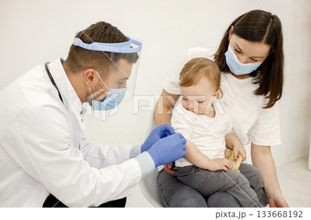 Male doctor in white medical gown is going to do vaccination. Boys and woman wearing white t-shirts. Doctor and mother wearing face masks. Male doctor in white medical gown is going to do vaccination. Boys and woman wearing white t-shirts. Doctor and mother wearing face masks. 133668192